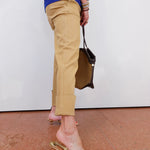 Person wearing beige pants and gold sandals, holding a brown bag on a white background