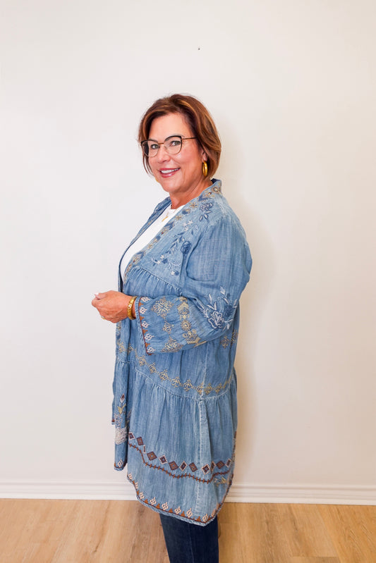 Woman wearing a blue embroidered denim jacket against a white wall.