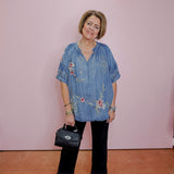 Woman wearing a blue embroidered blouse with black pants and shoes, holding a black handbag against a pink wall.