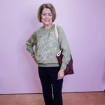 Woman wearing a green sweater with text and graphics, holding a brown bag against a light purple wall.