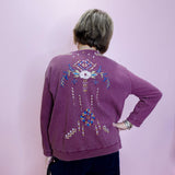 Person wearing a purple sweater with decorative patterns on the back, standing against a plain background.