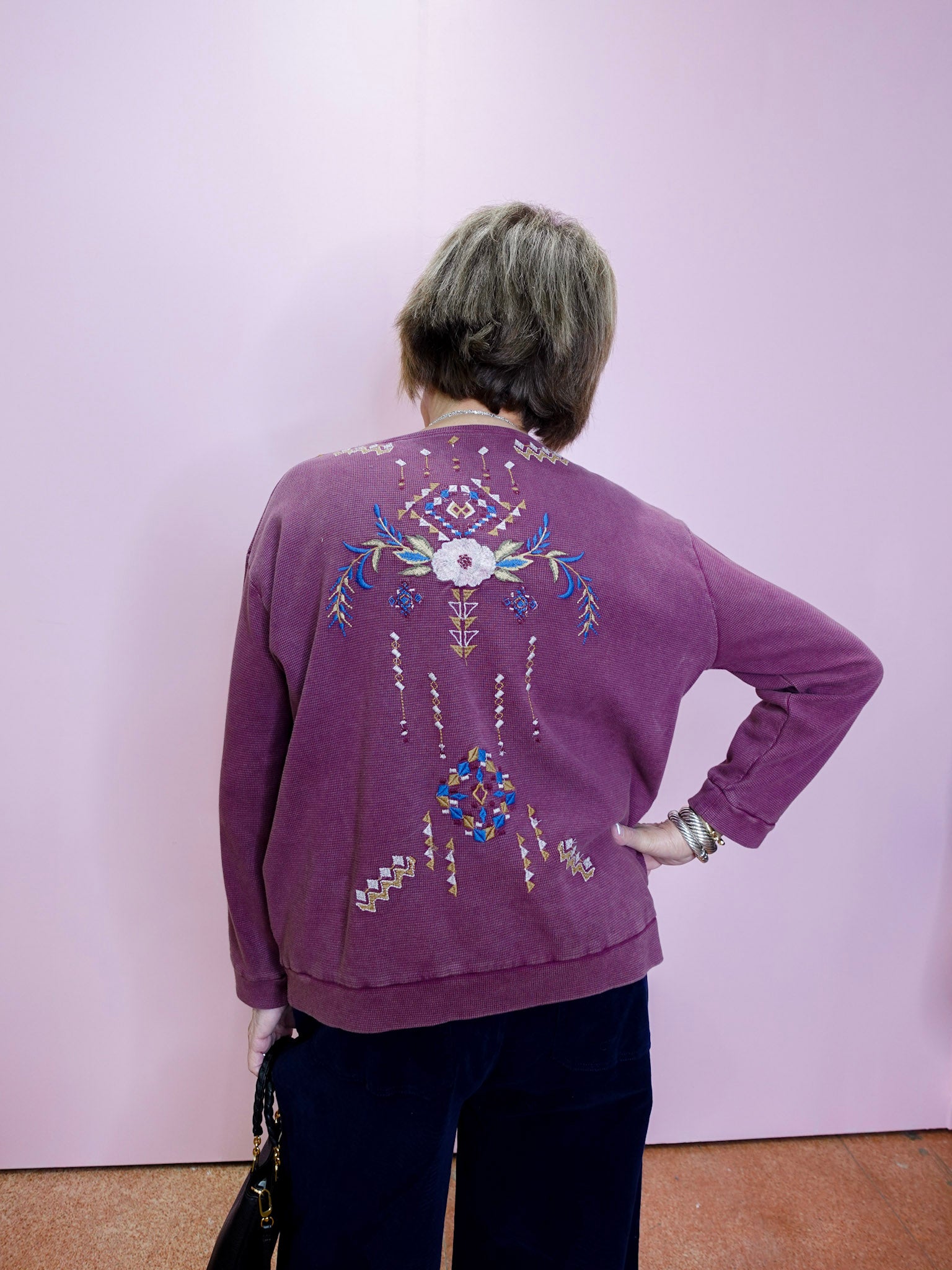 Person wearing a purple sweater with decorative patterns on the back, standing against a plain background.