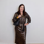 Woman wearing a leopard print dress with a black handbag against a white wall.