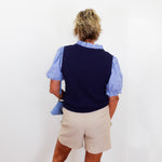 Person wearing a navy vest over a striped shirt with beige shorts, standing against a white wall.