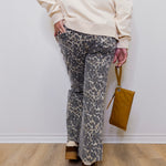 Person wearing a beige sweater and patterned pants holding a brown bag against a white wall.