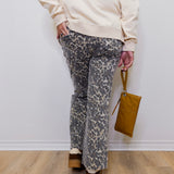 Person wearing a beige sweater and patterned pants holding a brown bag against a white wall.