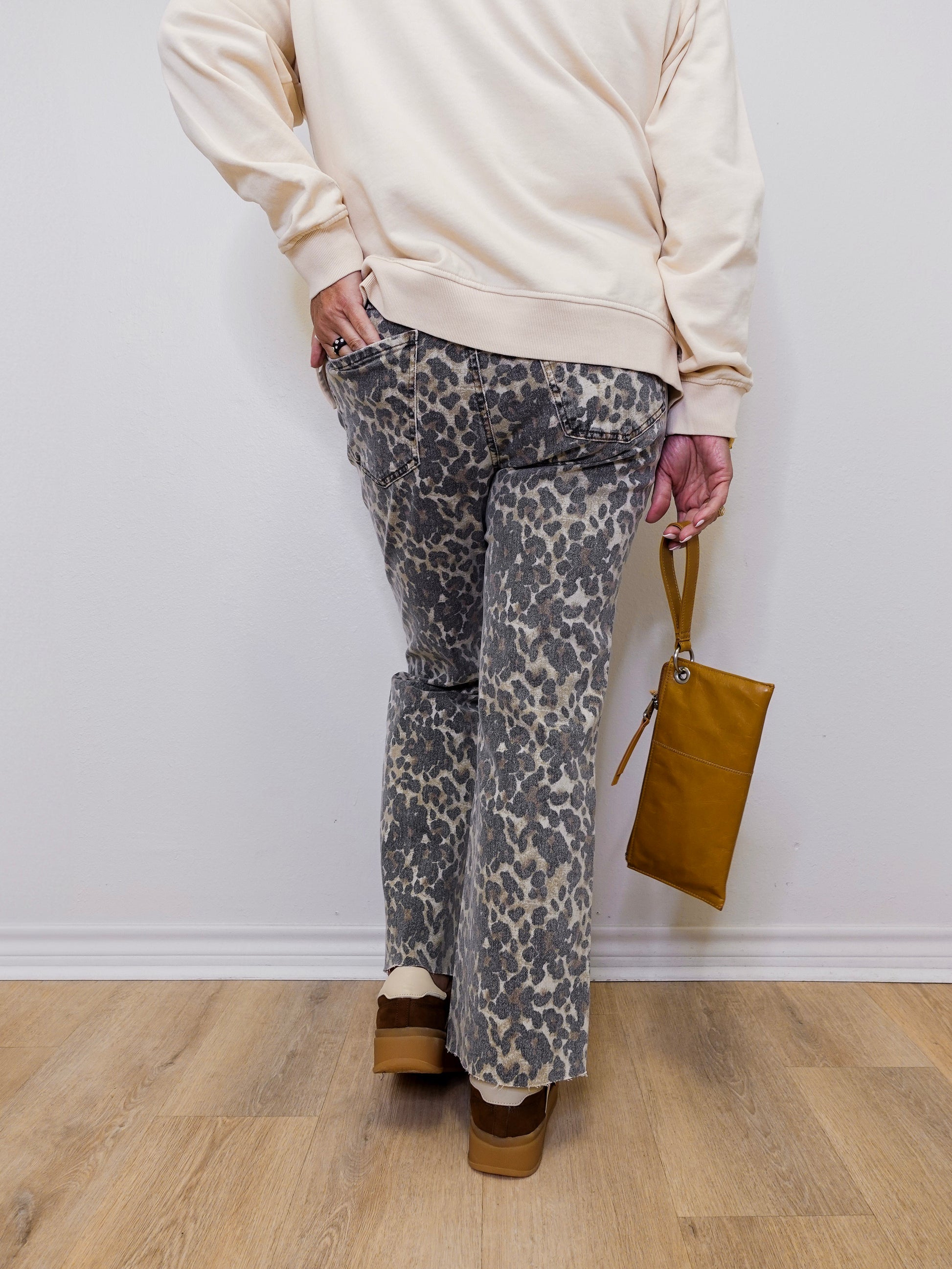 Person wearing a beige sweater and patterned pants holding a brown bag against a white wall.