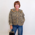 Woman wearing a leopard print jacket and blue jeans, holding a black handbag against a white wall.