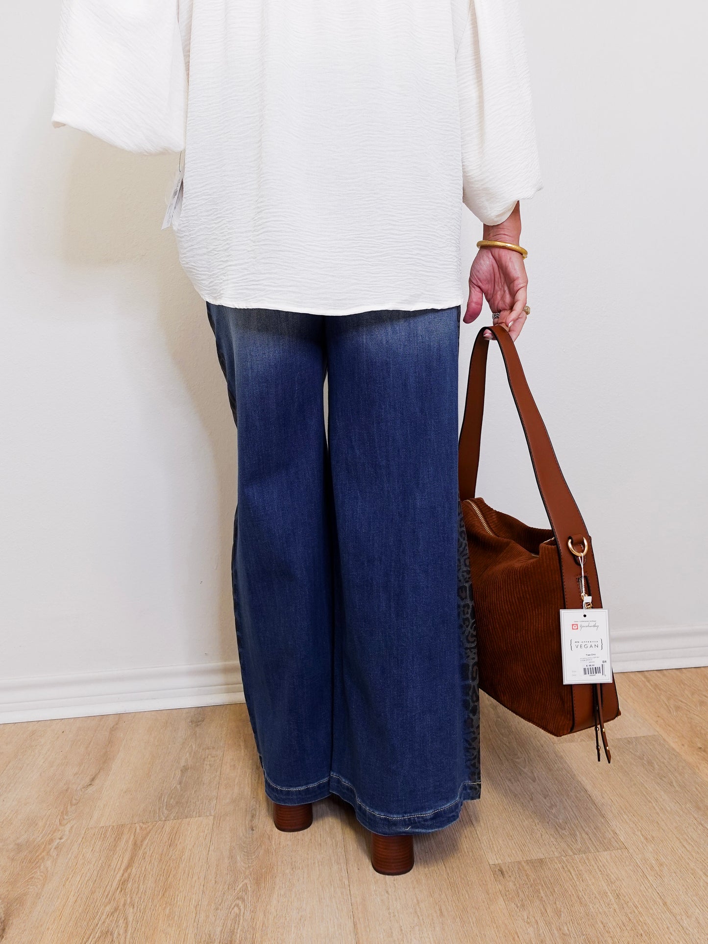 Person wearing blue jeans and a white top, holding a brown handbag with a tag.