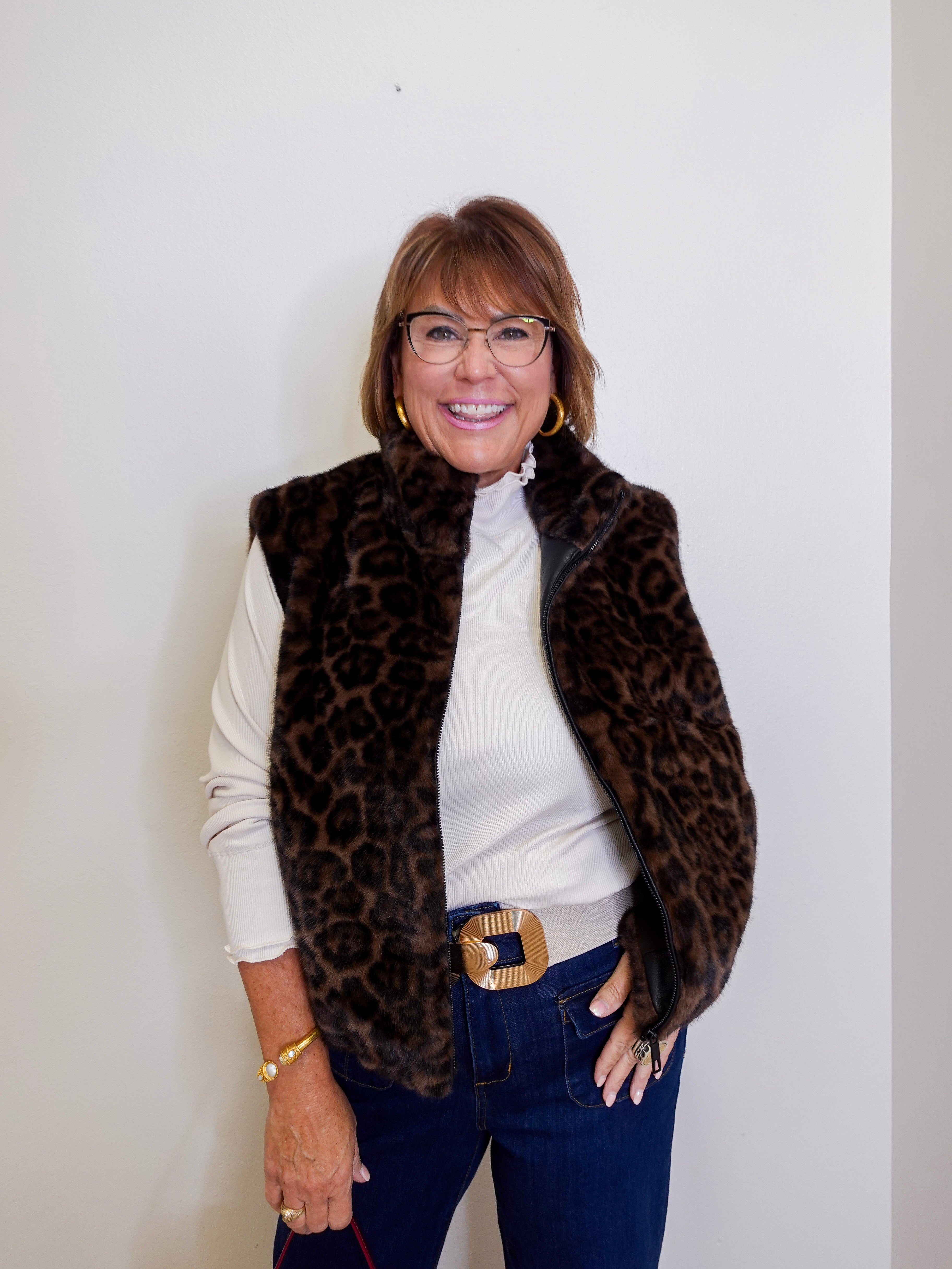 Woman wearing a leopard print vest over a white shirt with blue jeans against a plain background