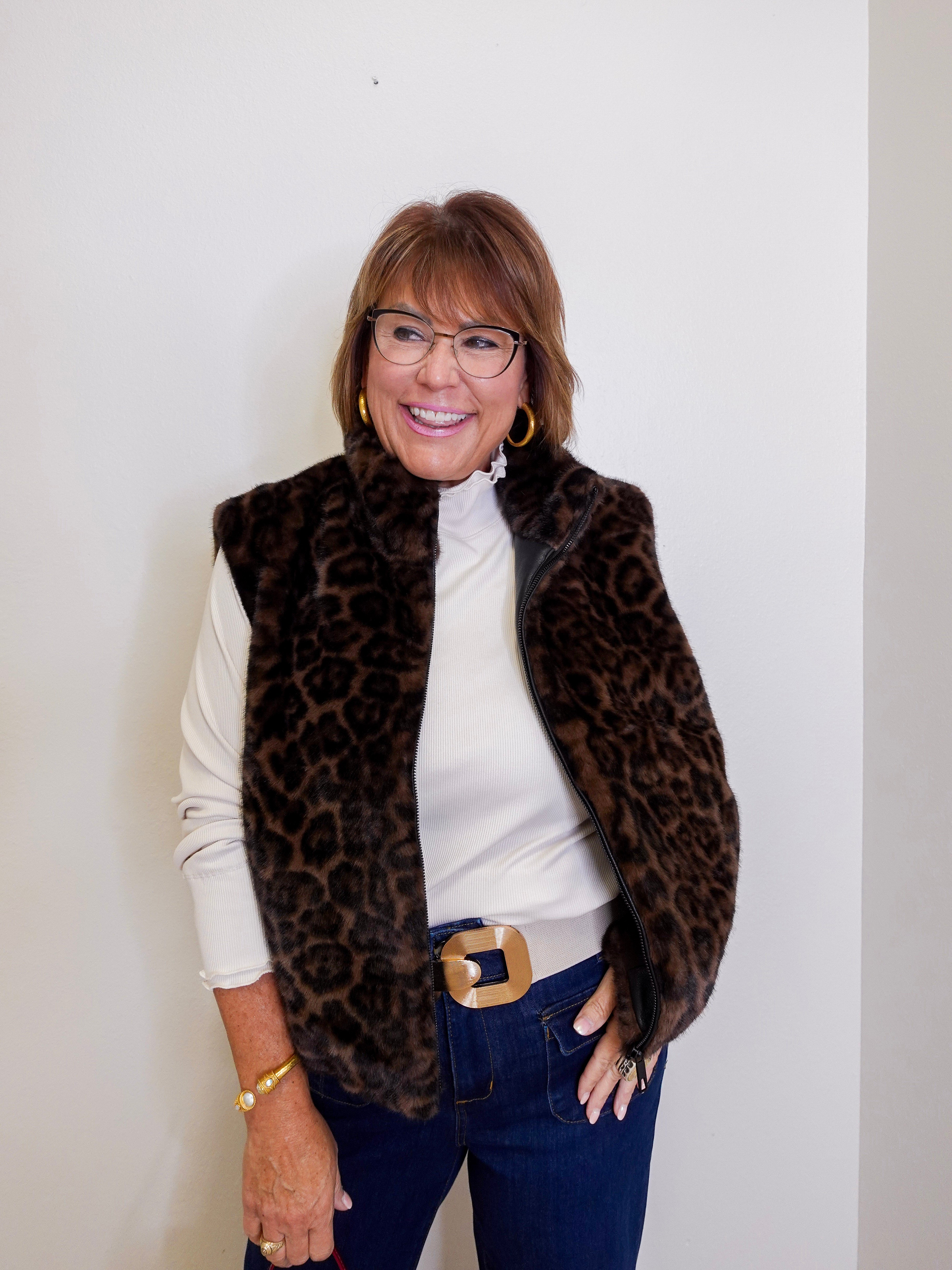 Woman wearing a leopard print vest over a white shirt with blue jeans against a plain background