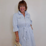 Woman wearing a light blue dress with a belt, standing against a plain wall.
