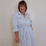 Woman wearing a light blue dress with a belt, standing against a plain wall.