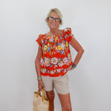 Woman wearing a colorful floral top and beige shorts, holding a straw bag against a white background.