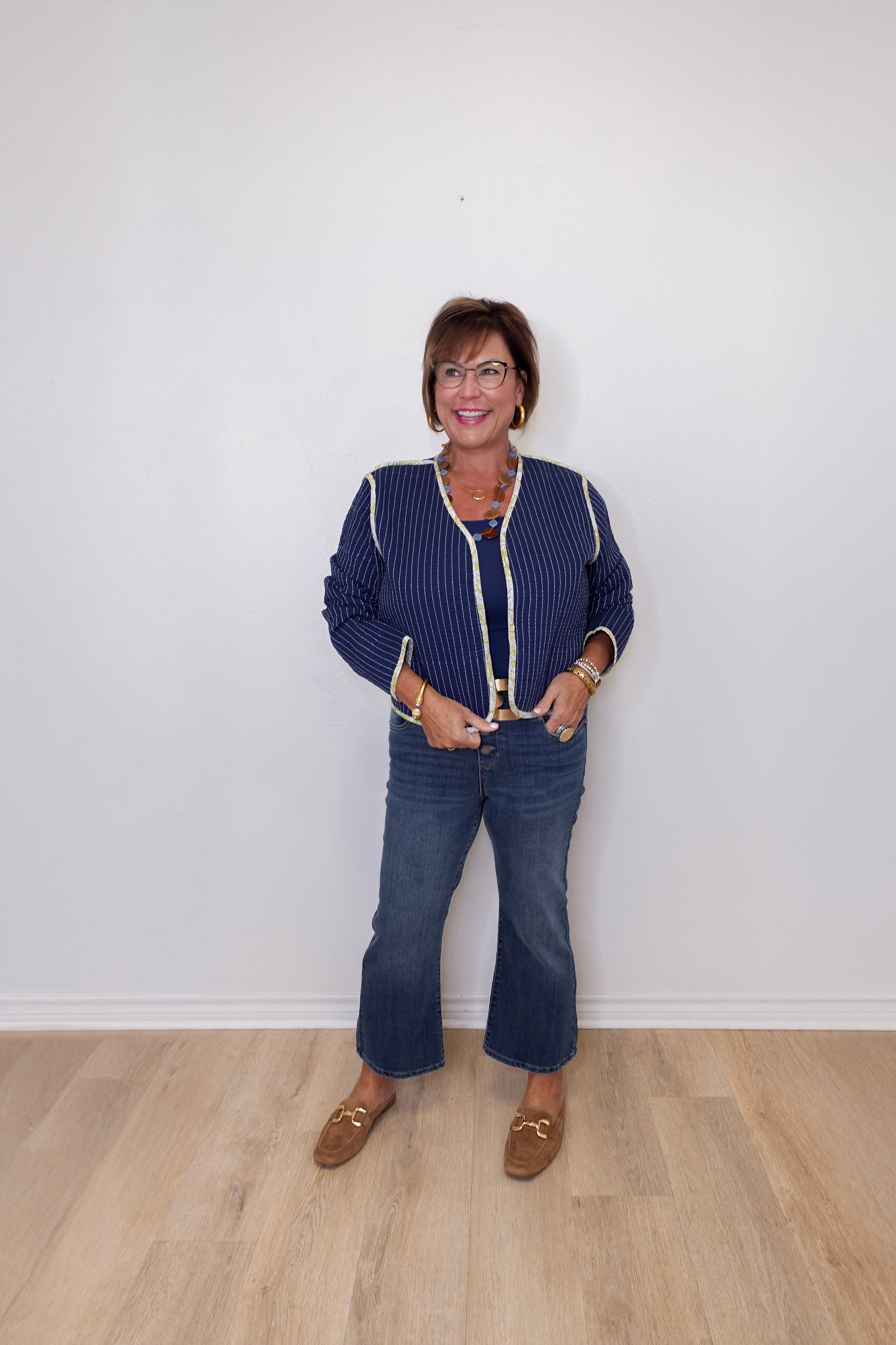 Woman wearing a blue patterned jacket and jeans standing against a white wall.