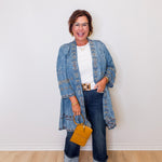 Woman wearing a denim jacket, white shirt, and jeans, holding a yellow handbag against a white wall.