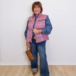Woman wearing a pink quilted vest over a denim jacket with blue jeans and brown boots, holding a brown bag against a white background.