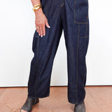A person wearing dark indigo wide-leg denim cargo pants with contrast yellow stitching, paired with a matching dark denim top and black snakeskin-embossed heeled sandals, standing against a white wall.