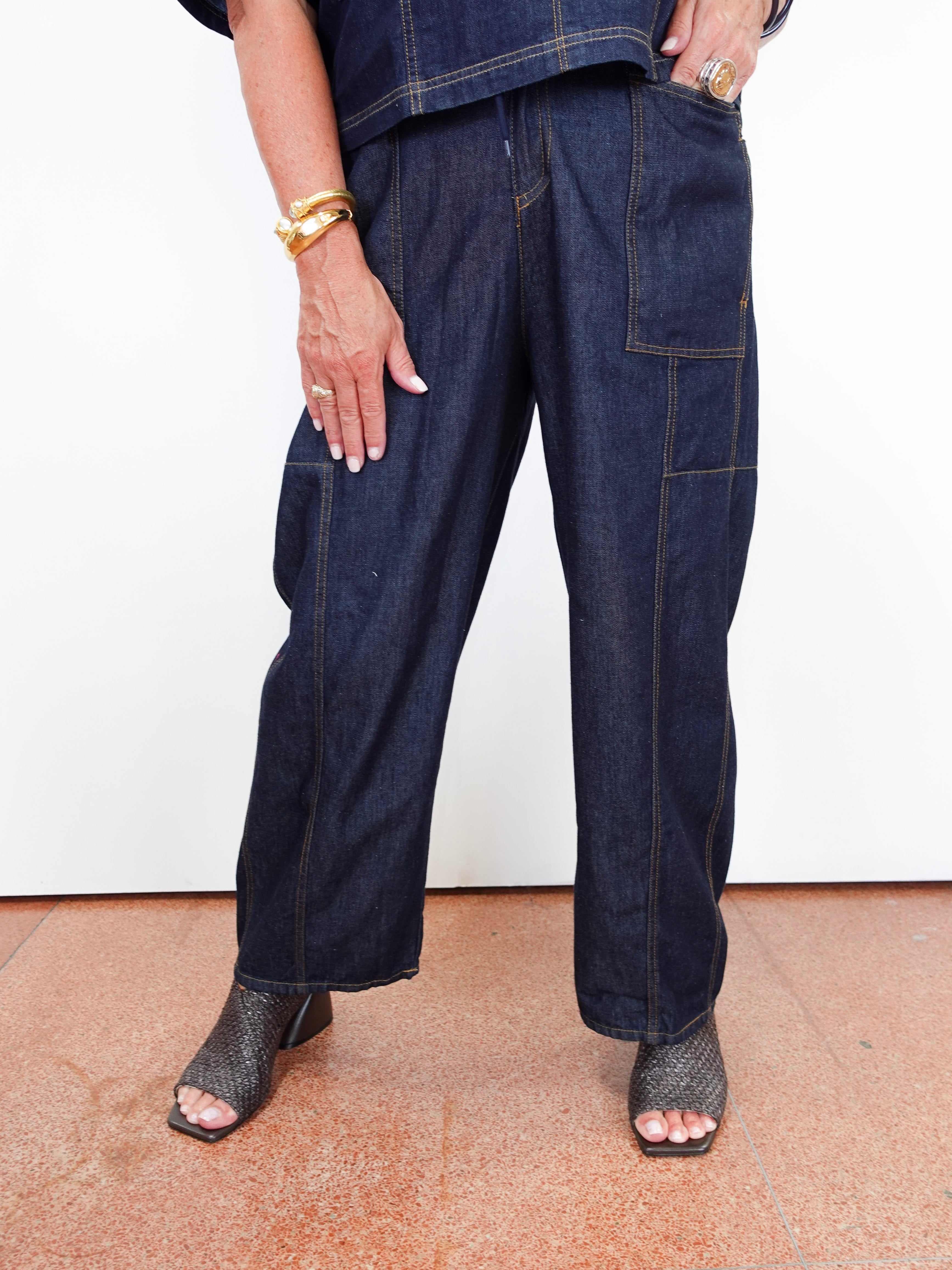 A person wearing dark indigo wide-leg denim cargo pants with contrast yellow stitching, paired with a matching dark denim top and black snakeskin-embossed heeled sandals, standing against a white wall.