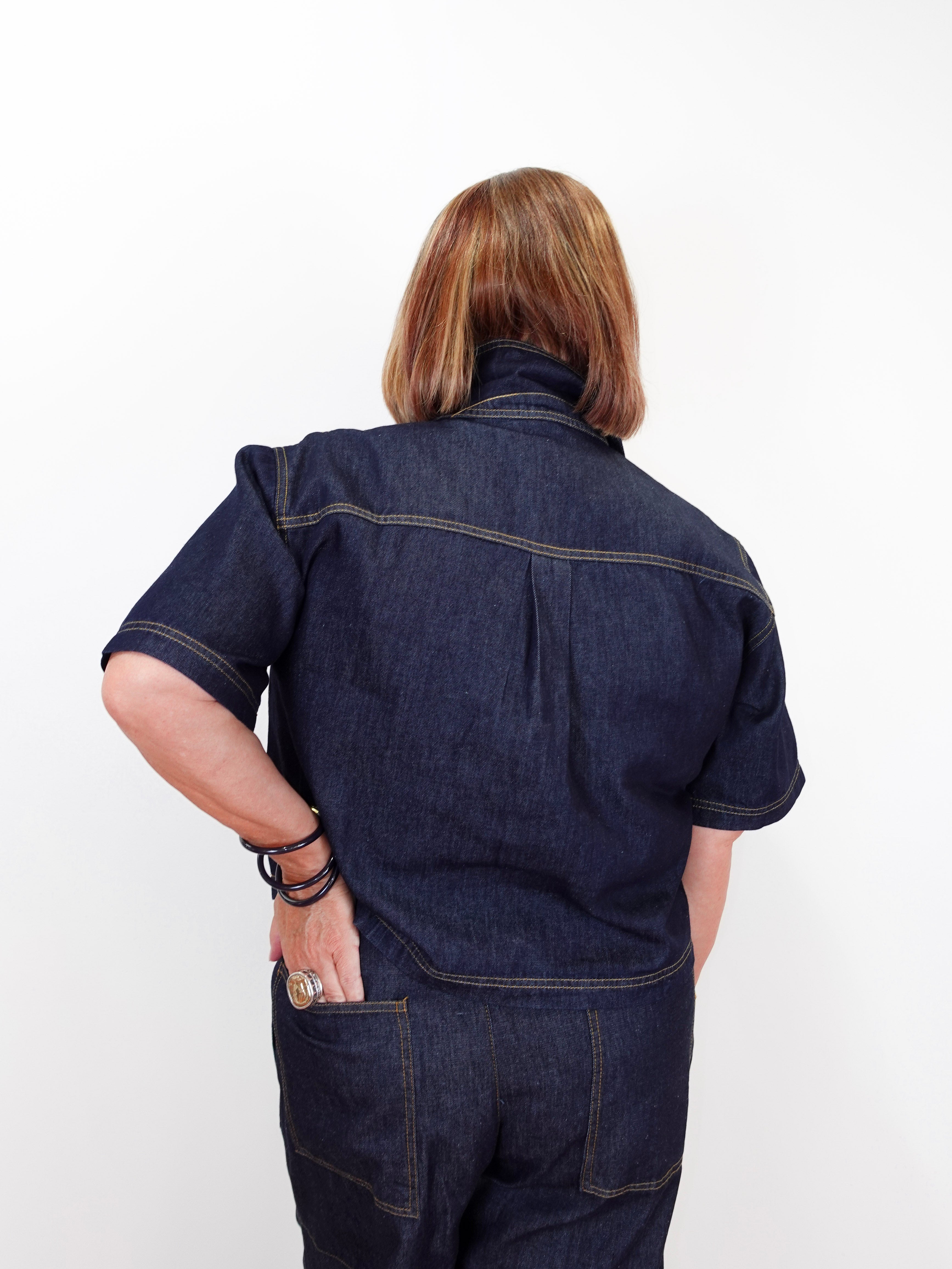 A person shown from the back wearing a dark indigo denim short-sleeve work shirt and matching denim pants against a plain white background.