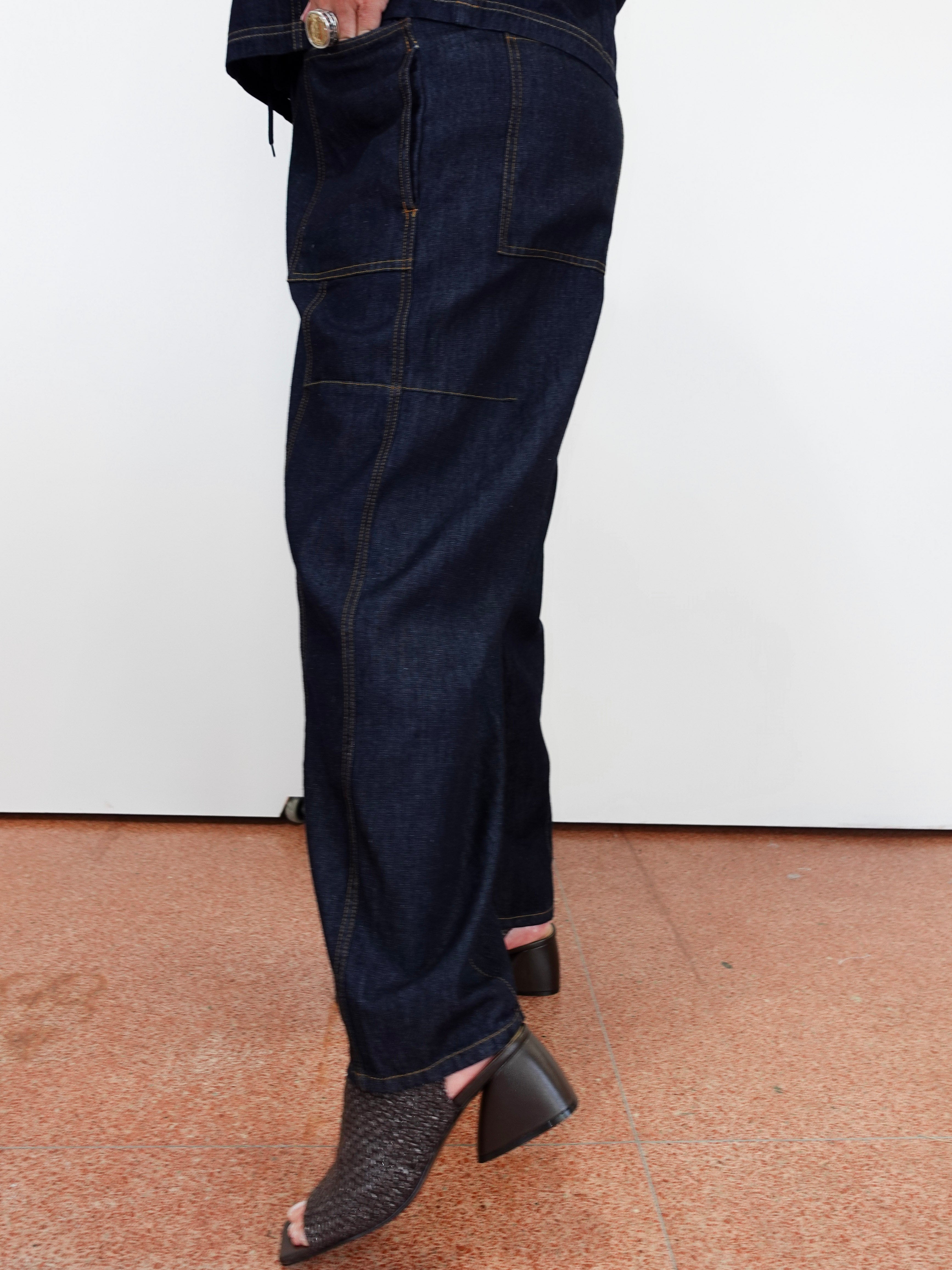 A person wearing dark indigo denim jeans with a relaxed straight-leg fit and black snakeskin-textured peep-toe block-heel mules, standing against a white wall on a terracotta tiled floor.