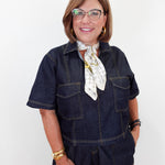 A smiling woman with glasses and a short bob haircut models a dark indigo denim short-sleeve jumpsuit with contrast stitching, styled with a white patterned neck scarf and gold jewelry against a plain white background.