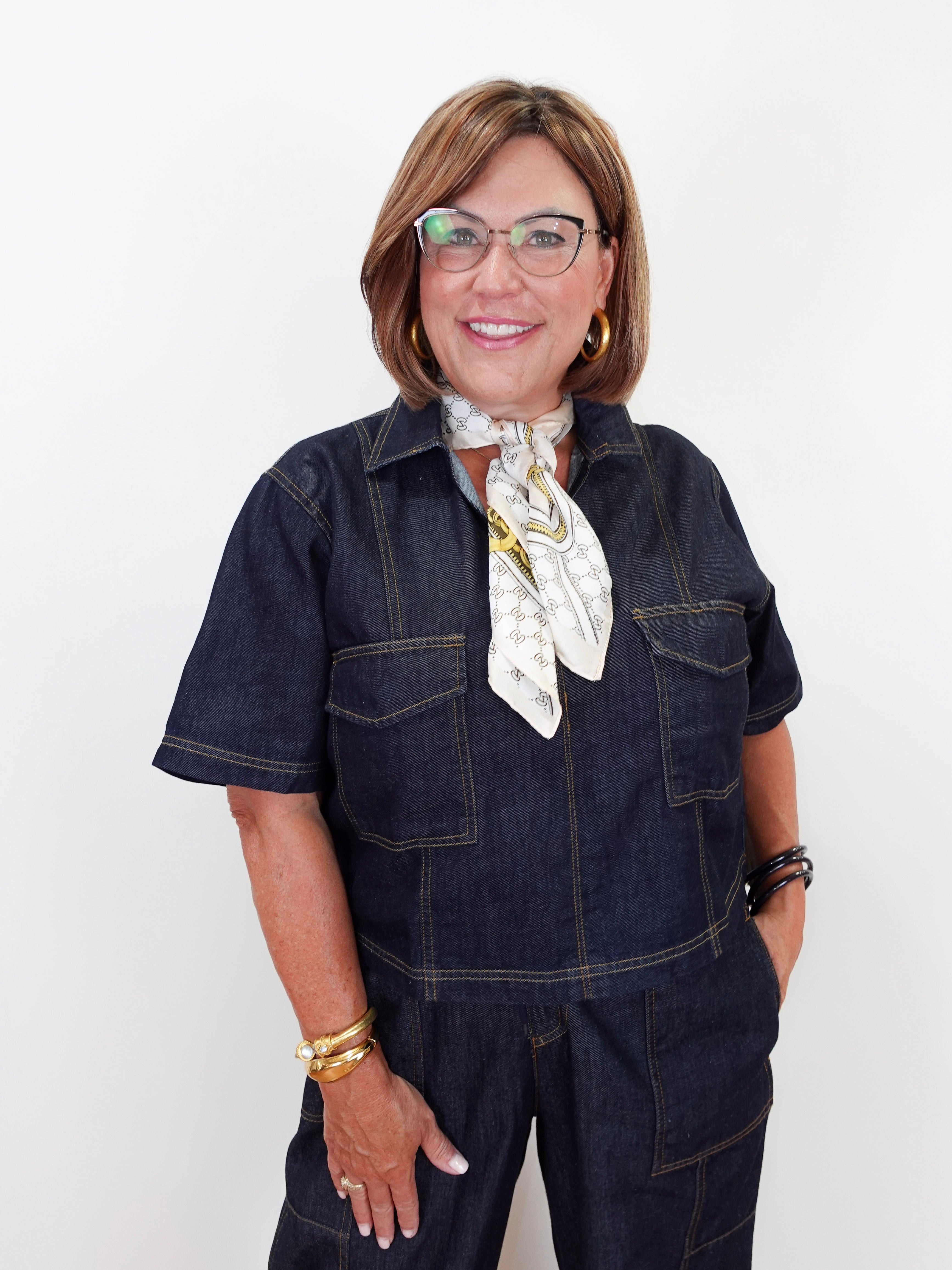 A smiling woman with glasses and a short bob haircut models a dark indigo denim short-sleeve jumpsuit with contrast stitching, styled with a white patterned neck scarf and gold jewelry against a plain white background.
