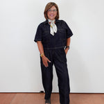A woman wearing dark indigo denim short-sleeve jumpsuit overalls with a white patterned neck scarf, standing against a plain white wall.