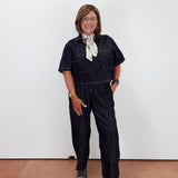 A woman wearing dark indigo denim short-sleeve jumpsuit overalls with a white patterned neck scarf, standing against a plain white wall.