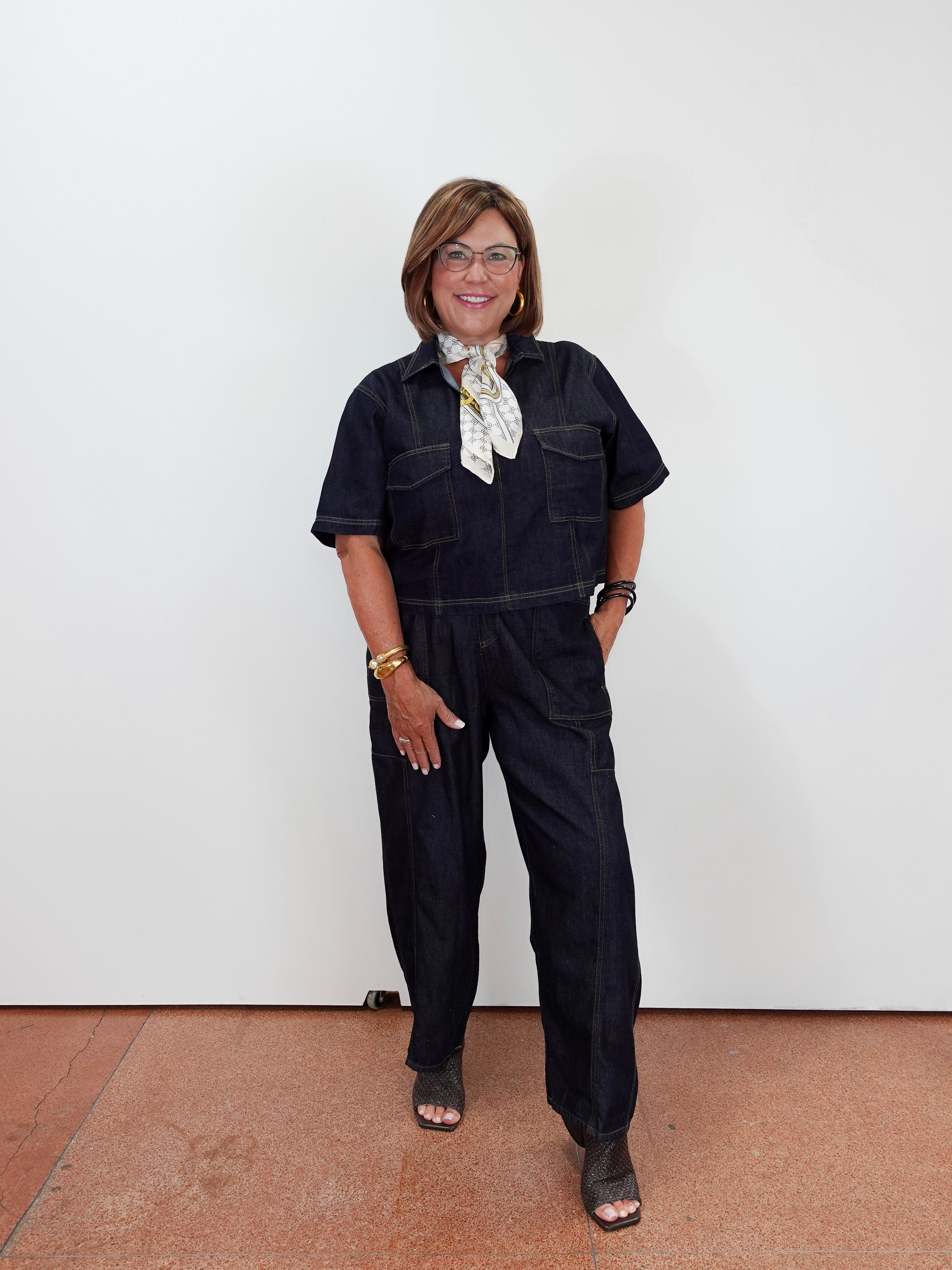 A woman wearing dark indigo denim short-sleeve jumpsuit overalls with a white patterned neck scarf, standing against a plain white wall.