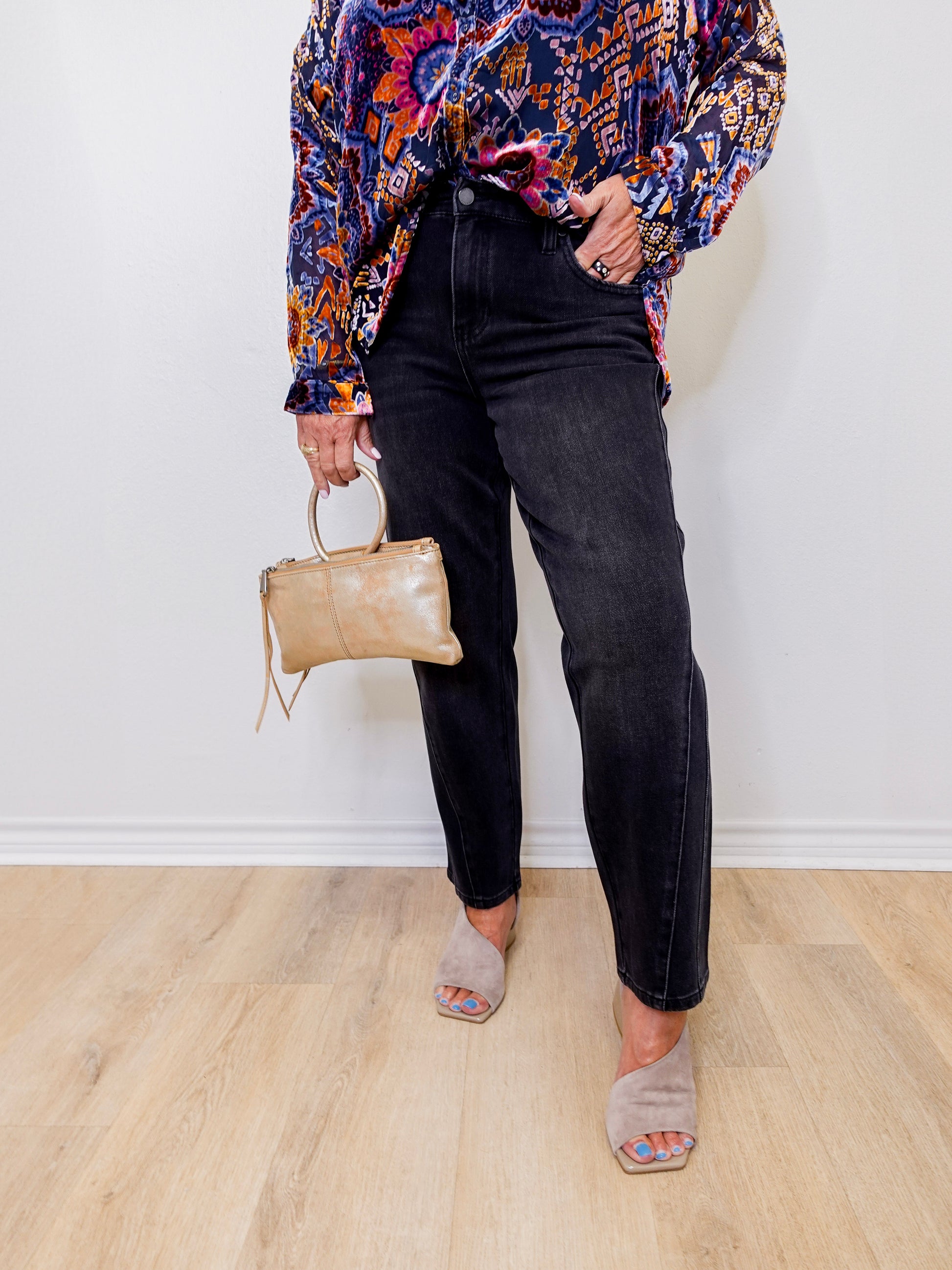 Person wearing a colorful patterned shirt, black jeans, and holding a beige handbag against a white wall.