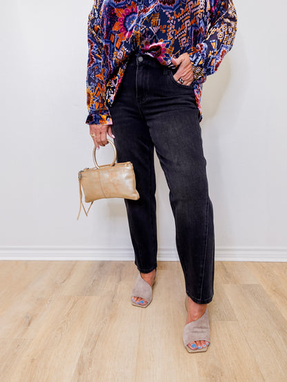Person wearing a colorful patterned shirt, black jeans, and holding a beige handbag against a white wall.