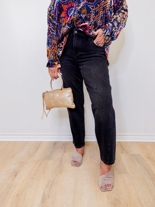 Person wearing a colorful patterned shirt, black jeans, and holding a beige handbag against a white wall.