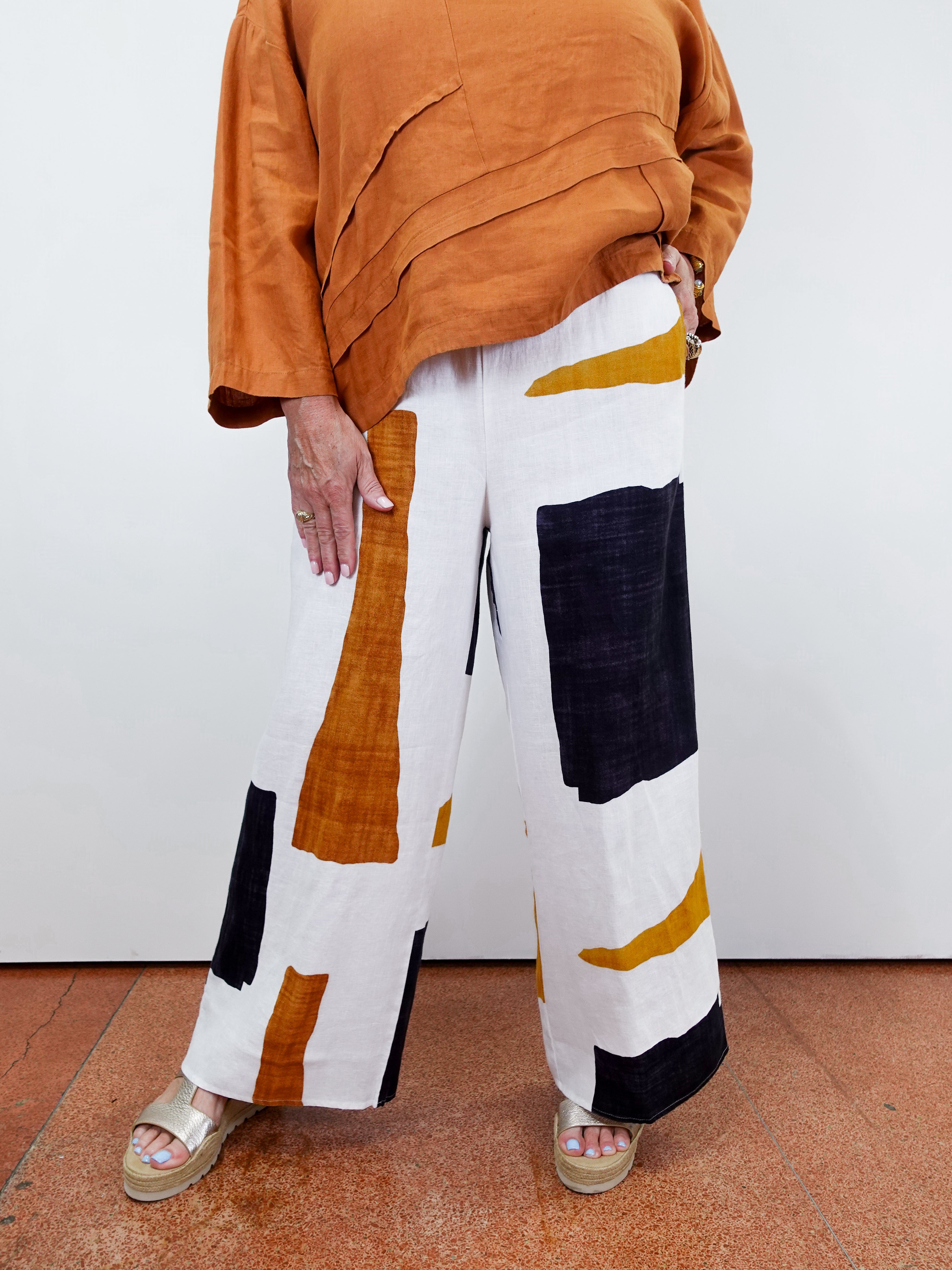 Person wearing a rust-colored top and white pants with geometric patterns on a plain background