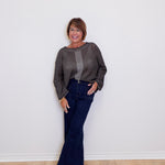 Woman wearing a gray sweater and blue jeans standing against a white wall.
