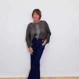 Woman wearing a gray sweater and blue jeans standing against a white wall.