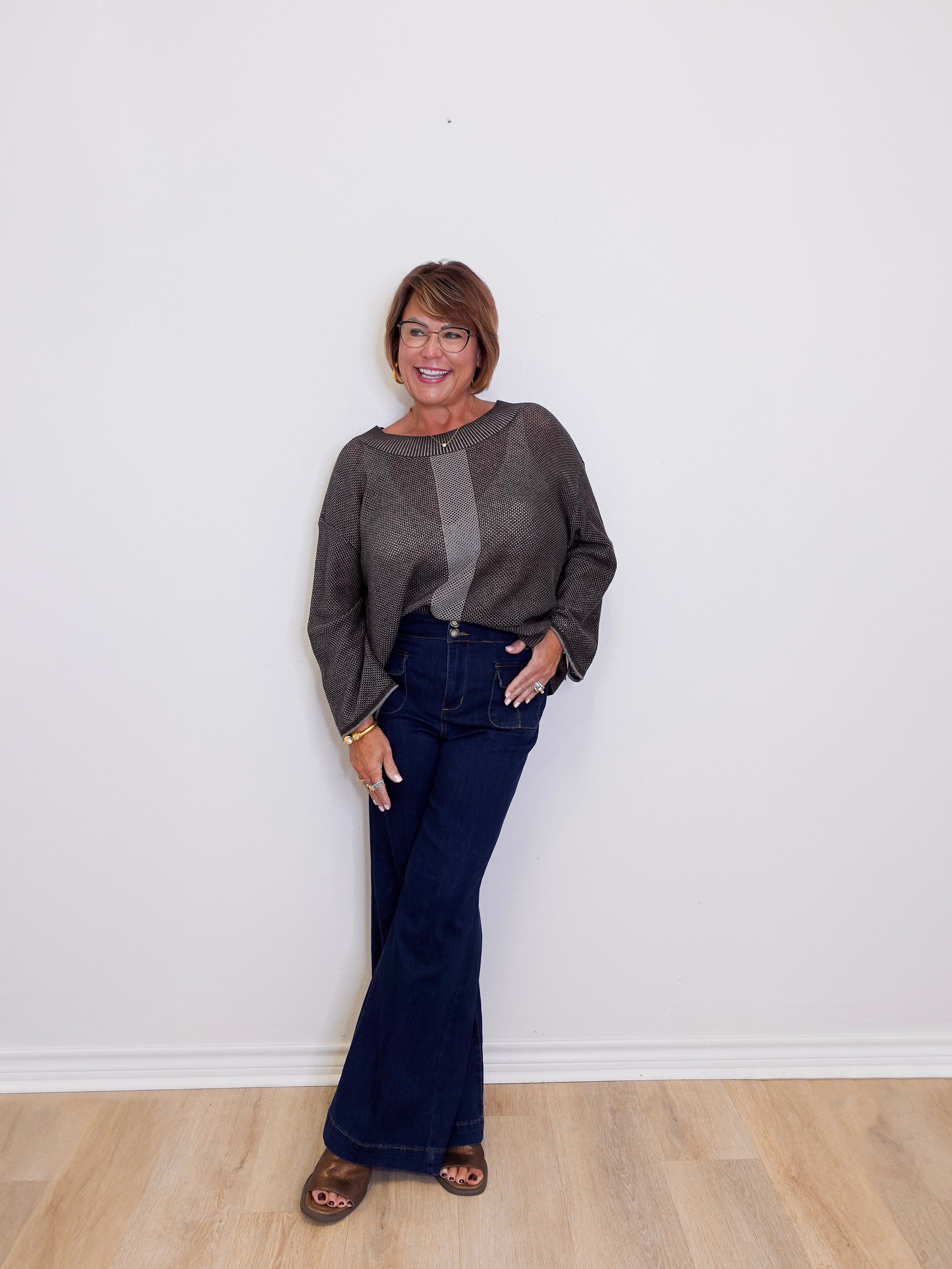 Woman wearing a gray sweater and blue jeans standing against a white wall.