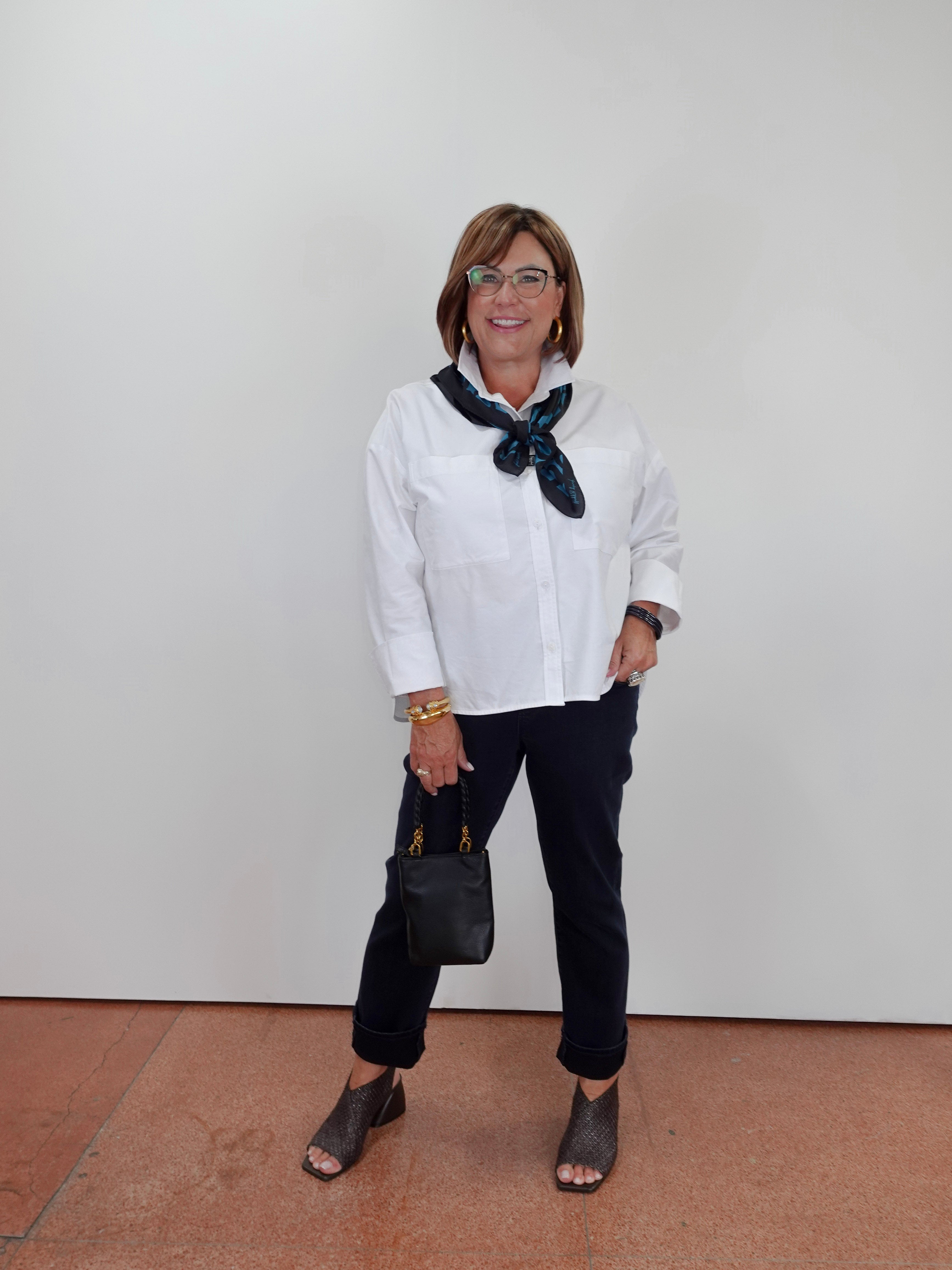 A woman modeling a white long-sleeve button-down shirt with a dark green patterned scarf, black pants, and dark gray peep-toe heels, standing against a plain white wall.