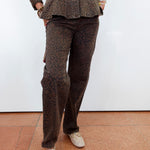 A person wearing a matching brown leopard-print jacket and trousers, paired with beige loafers, standing against a white wall on a terracotta tiled floor.