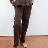 A person wearing a matching brown leopard-print jacket and trousers, paired with beige loafers, standing against a white wall on a terracotta tiled floor.