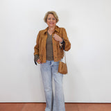 A woman stands against a plain white wall wearing a tan suede jacket over a black-and-white striped top, light-wash flare jeans, and black sandals, while carrying a tan crossbody bag.