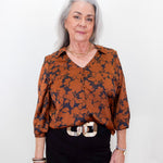 An older woman with gray hair stands against a plain white background wearing a rust-orange and navy floral patterned blouse, black trousers, and a chunky silver belt.