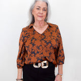 An older woman with gray hair stands against a plain white background wearing a rust-orange and navy floral patterned blouse, black trousers, and a chunky silver belt.
