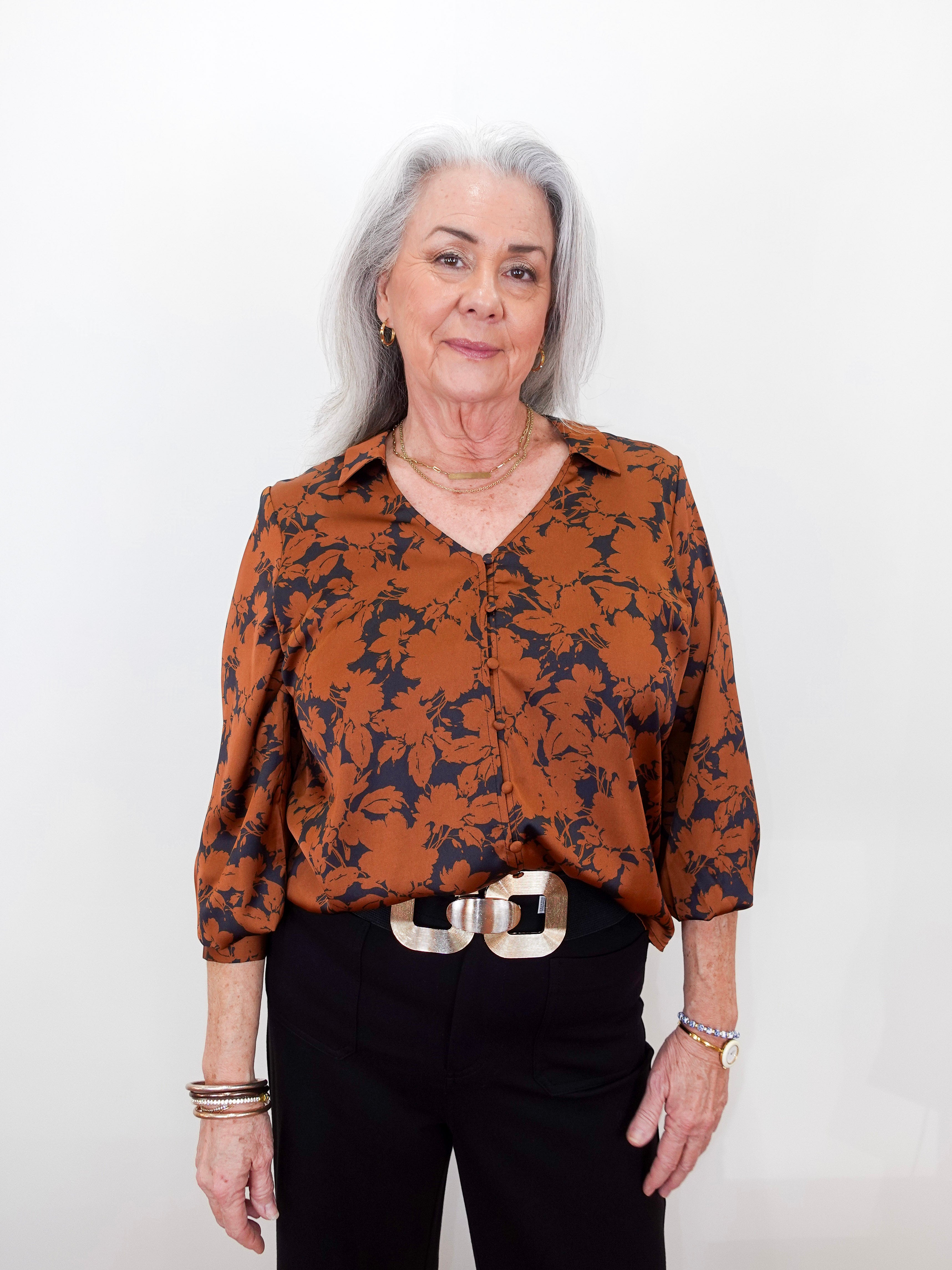 An older woman with gray hair stands against a plain white background wearing a rust-orange and navy floral patterned blouse, black trousers, and a chunky silver belt.
