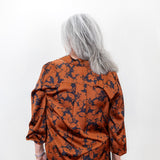 A woman with shoulder-length gray hair is shown from the back, wearing a long-sleeve satin blouse with a dark navy floral pattern over a rust-orange base, paired with black pants, against a plain white background.