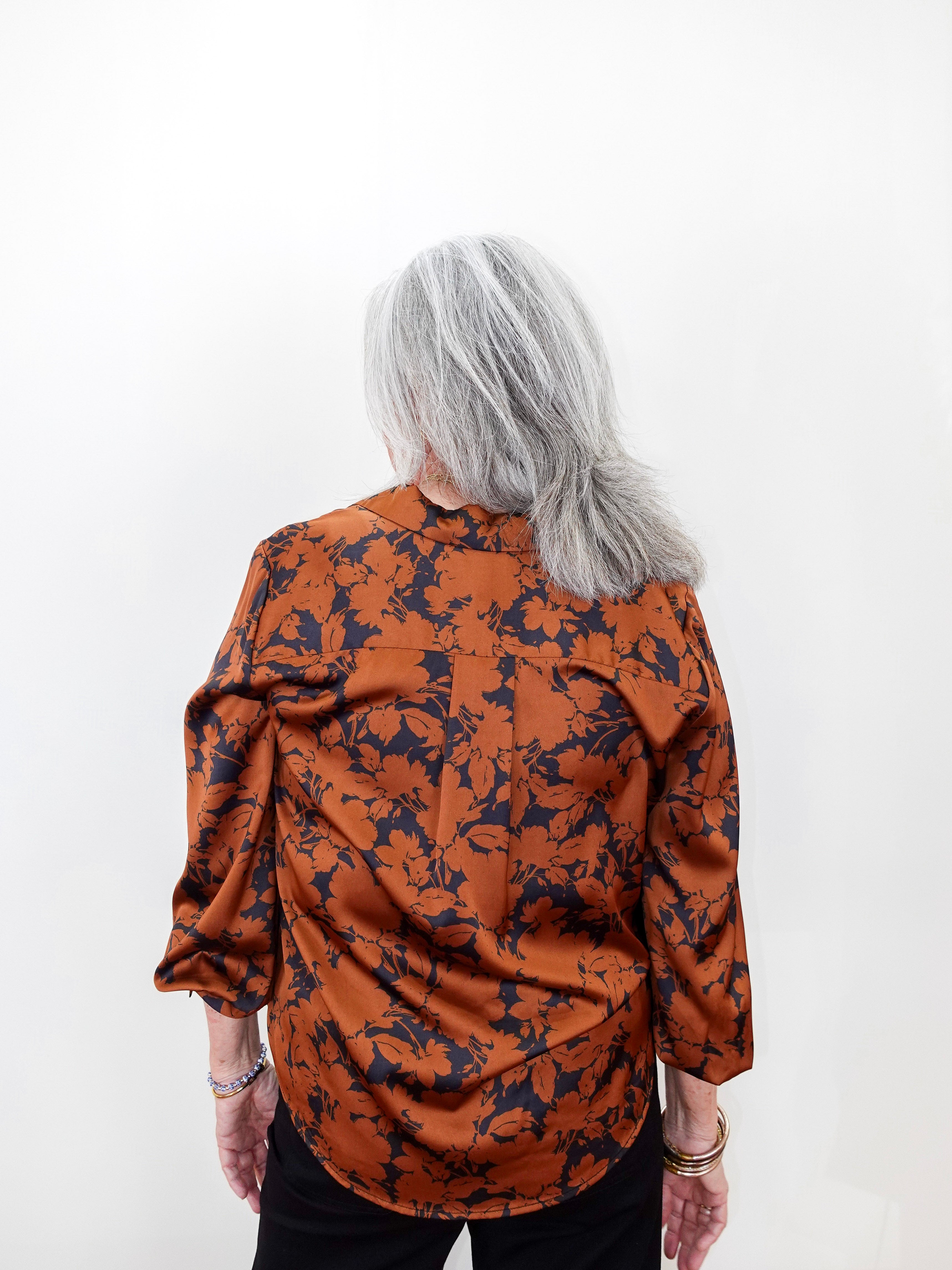 A woman with shoulder-length gray hair is shown from the back, wearing a long-sleeve satin blouse with a dark navy floral pattern over a rust-orange base, paired with black pants, against a plain white background.