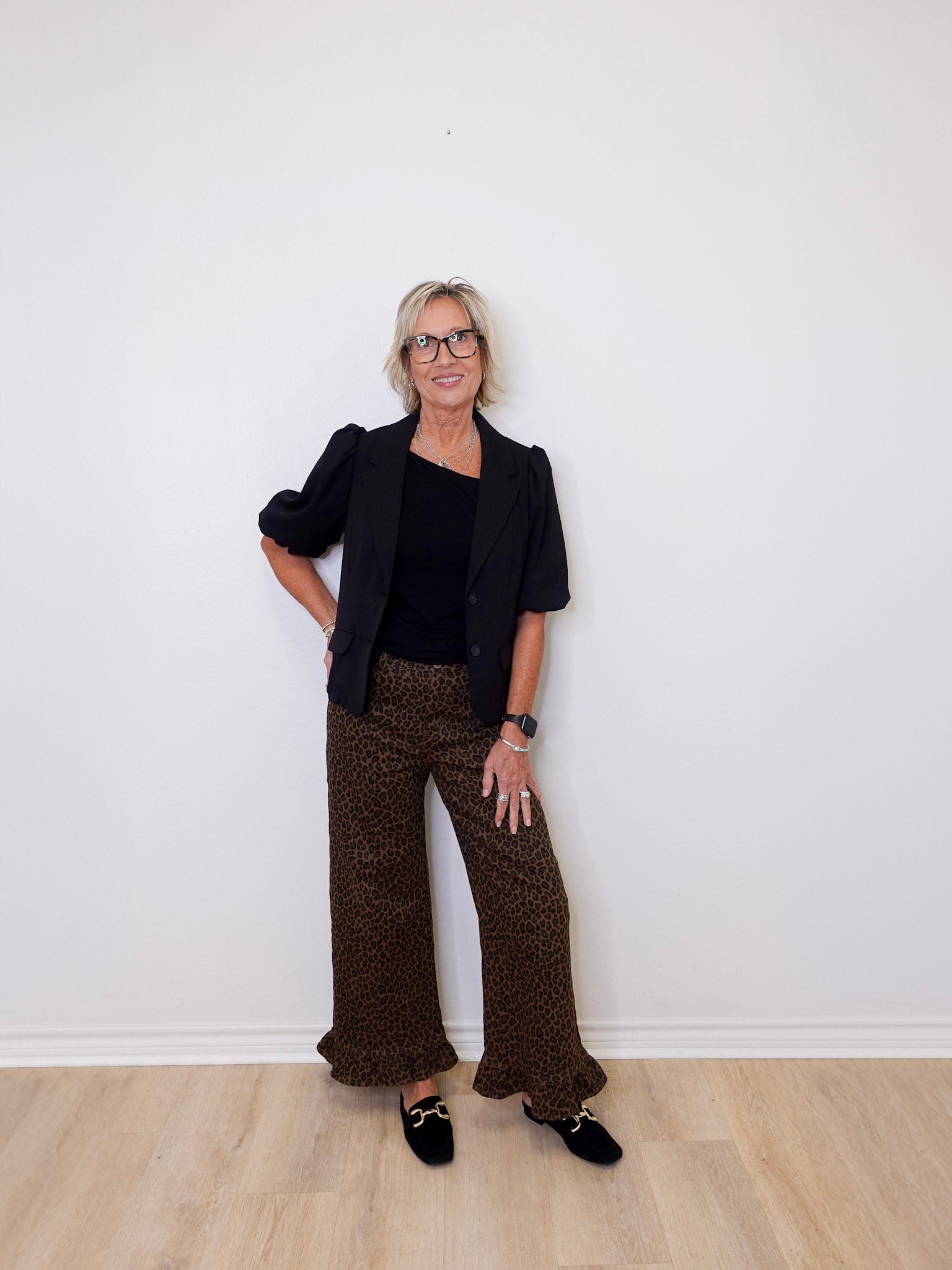 Woman wearing a black blazer and brown patterned pants against a white wall.