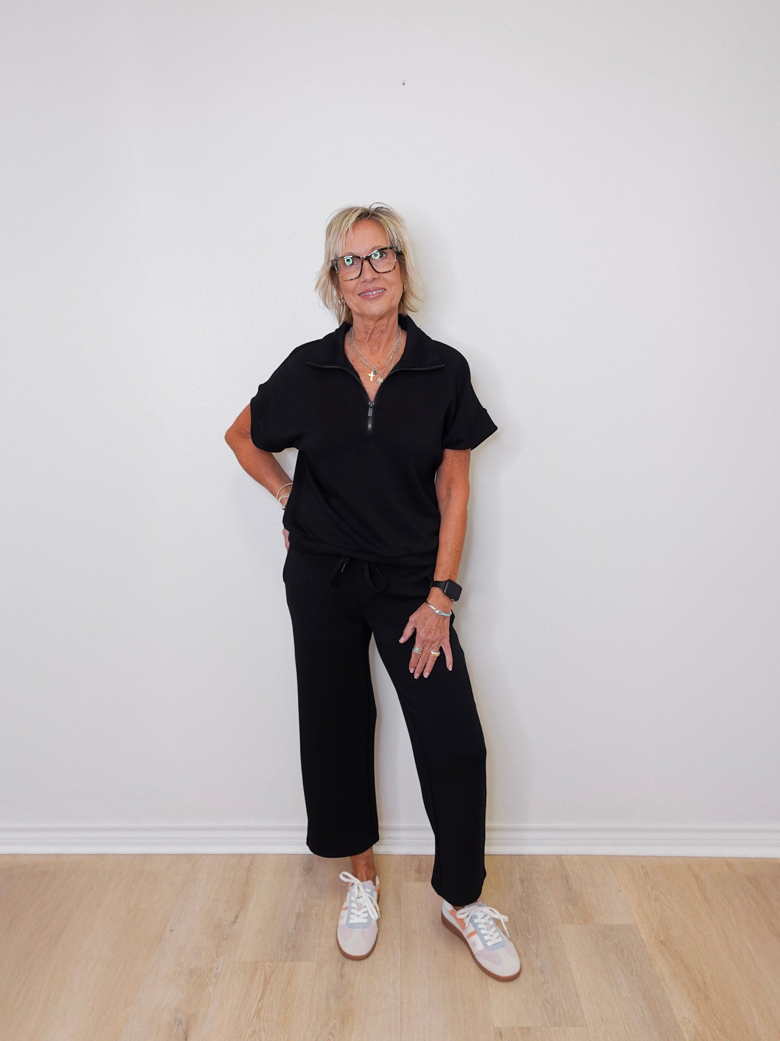 Person wearing a black outfit with white sneakers against a white wall