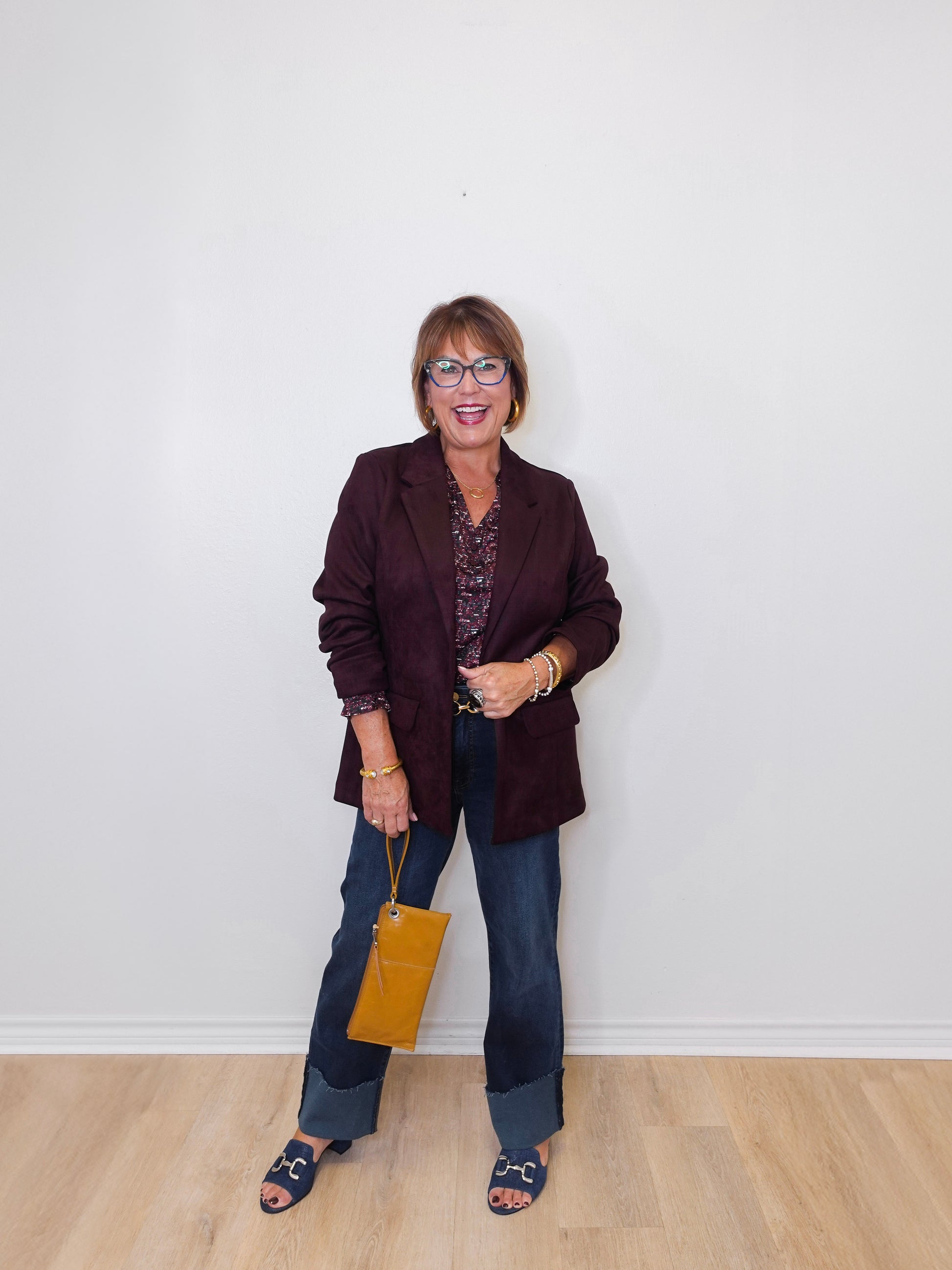 Woman wearing a dark blazer and jeans, holding a yellow handbag against a white wall.