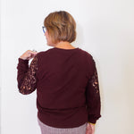 Person wearing a maroon sweater with lace sleeves against a white wall